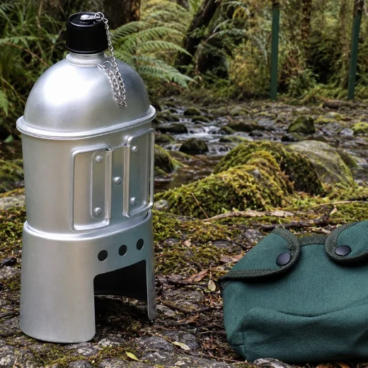 Assembled canteen stove with silver canteen and green carry bag beside a forest stream